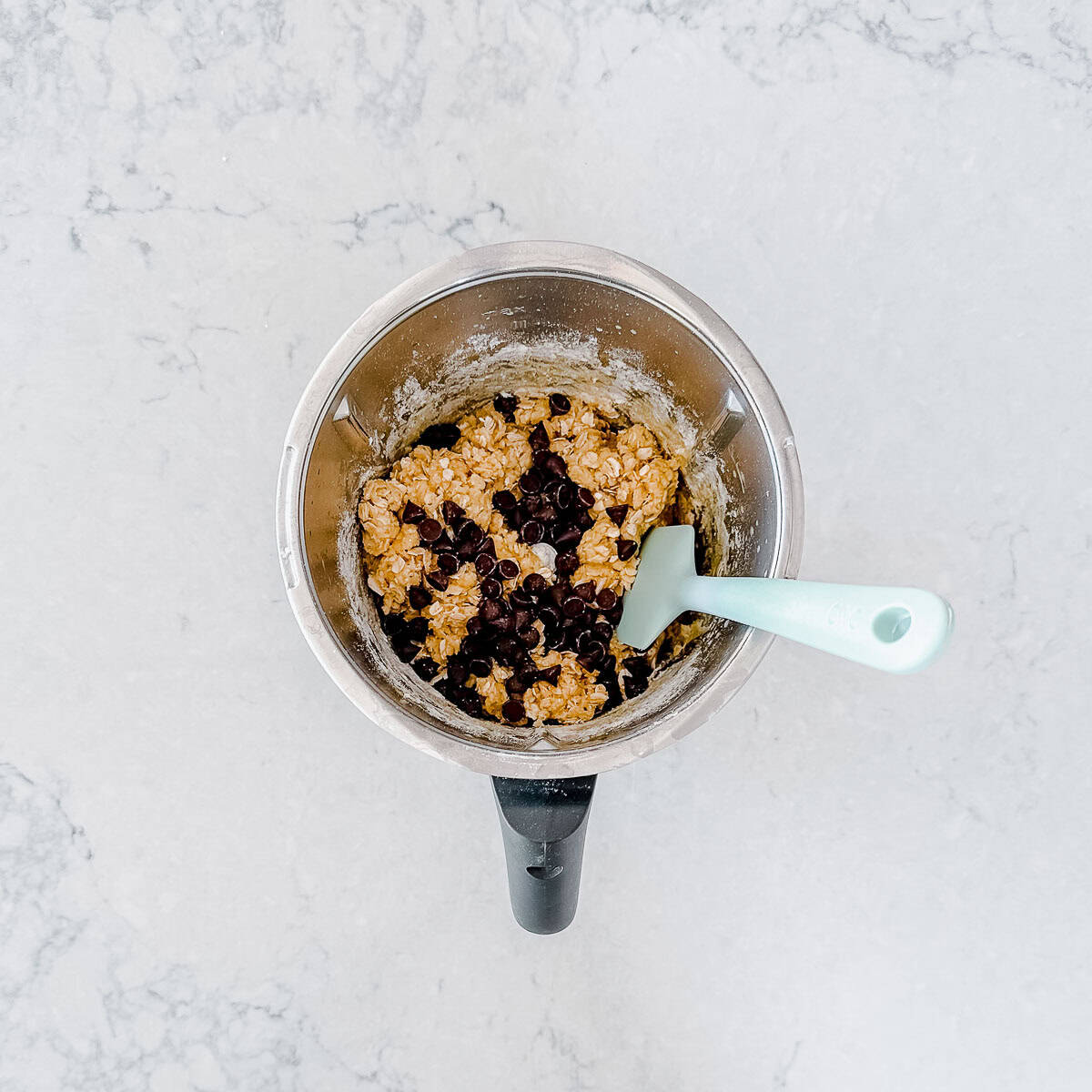 A Thermomix bowl with banana oat dough and chocolate chips, stirred with a light blue spatula, sits on a light marbled worktop.