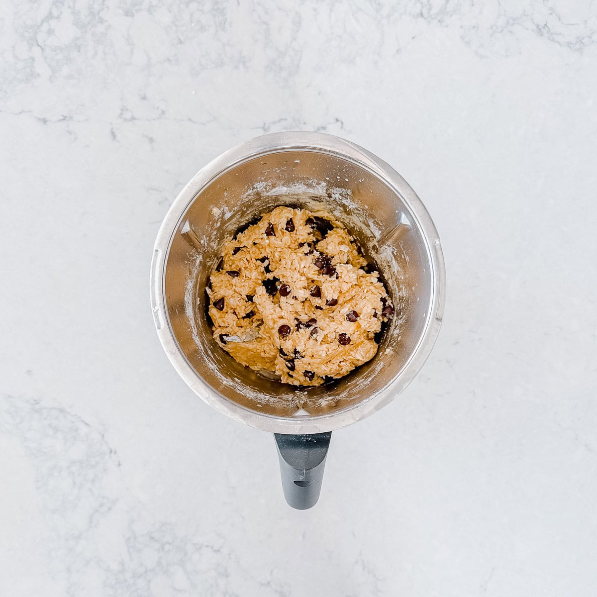 A mixing bowl with chocolate chip biscuit dough sits on a light marble worktop, seen from above.