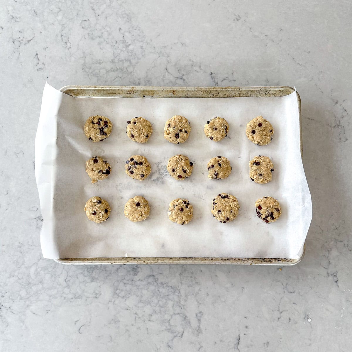 A baking tray lined with greaseproof paper holds fifteen evenly spaced balls of biscuit dough mixed with chocolate chips, ready to be baked, on a light grey countertop.