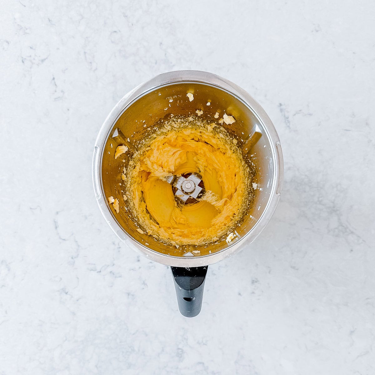 A food processor with a metal bowl contains a smooth orange mixture, likely puréed pumpkin, seen from above on a white marble worktop.