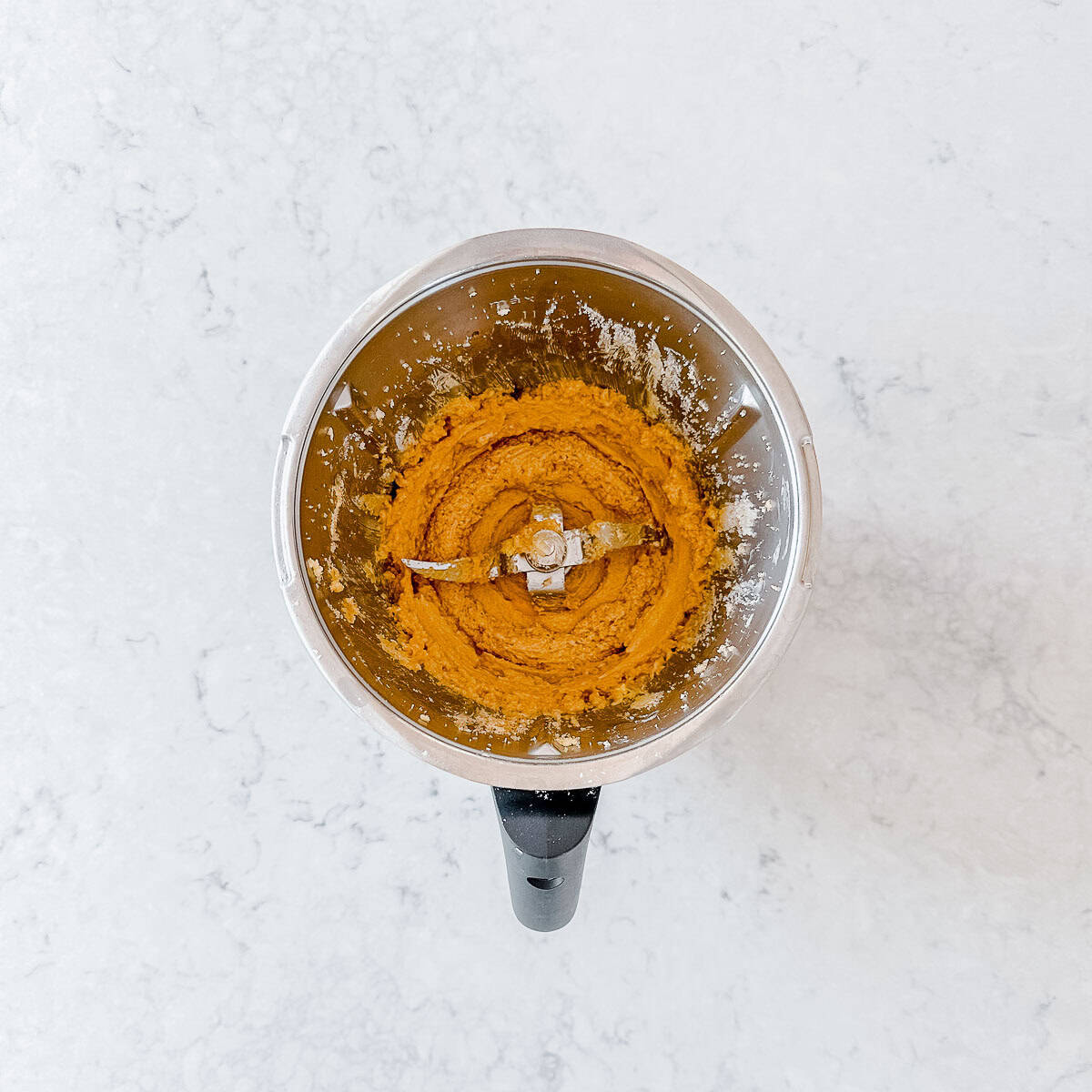 A food processor bowl with cookie dough mixture viewed from above on a white marble surface.