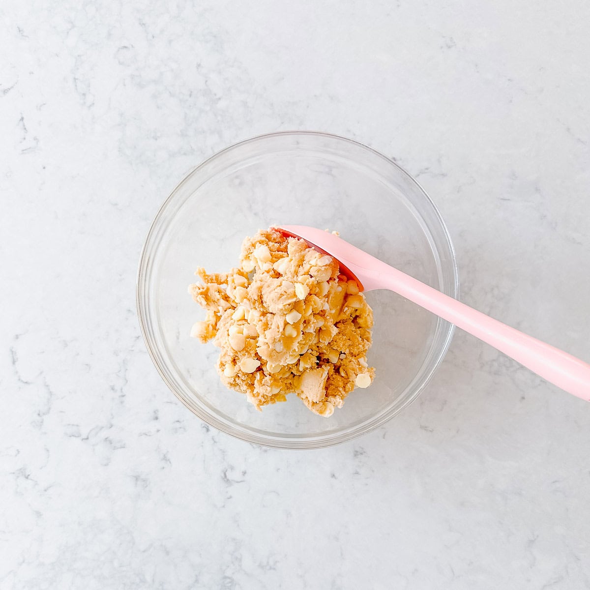A glass bowl with crumbled dough or biscuit mixture and a pink spatula on a light, marbled surface.