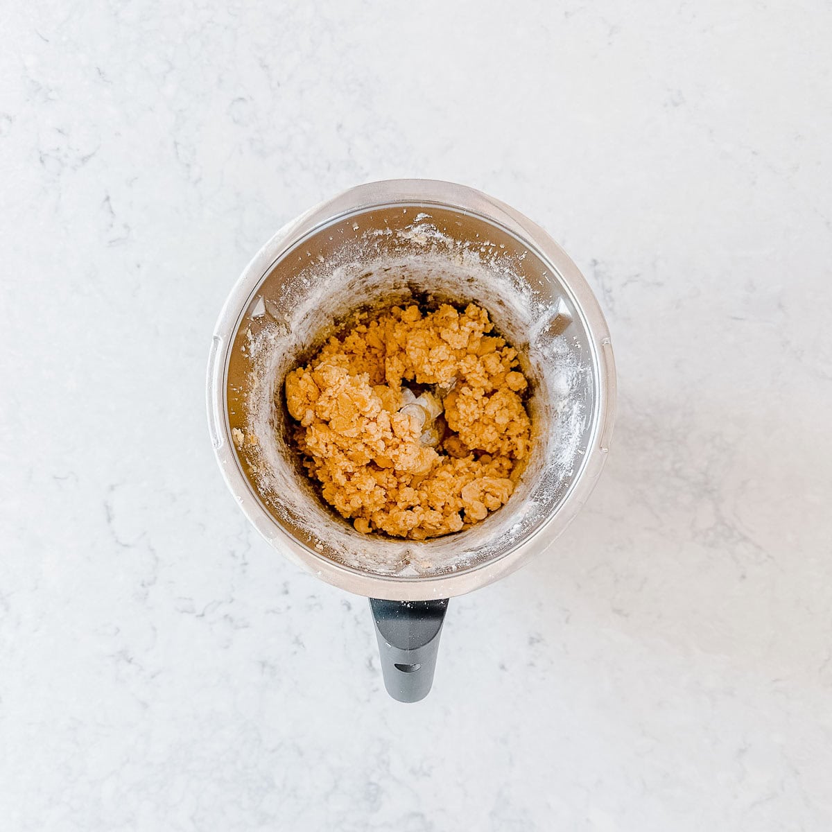 A metal mixing bowl contains a mound of light brown biscuit dough, partially mixed, sitting on a white marble worktop.