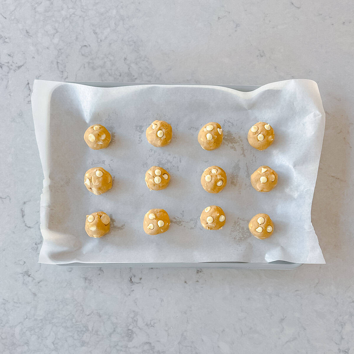 A baking tray lined with baking paper holds 14 evenly spaced balls of cookie dough with white chocolate chips, ready to be baked.