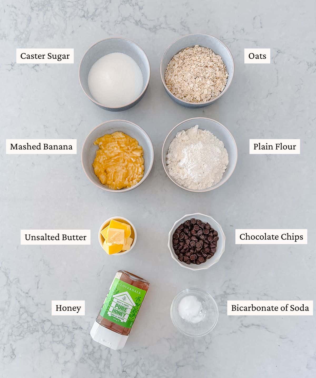 Overhead view of labelled bowls containing caster sugar, oats, mashed banana, plain flour, unsalted butter, chocolate chips, honey, and bicarbonate of soda on a light worktop.
