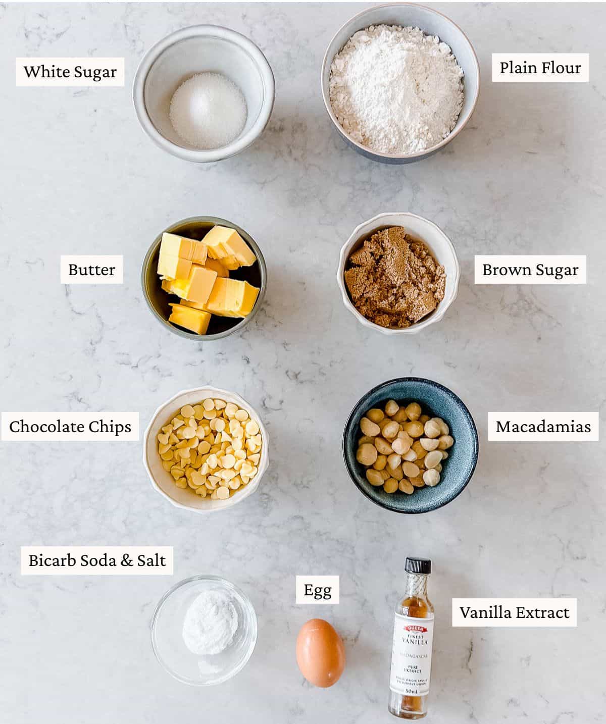 Top-down view of labelled baking ingredients in bowls: white sugar, plain flour, butter, brown sugar, chocolate chips, macadamias, bicarb soda & salt, egg, and a bottle of vanilla extract on a marble surface.