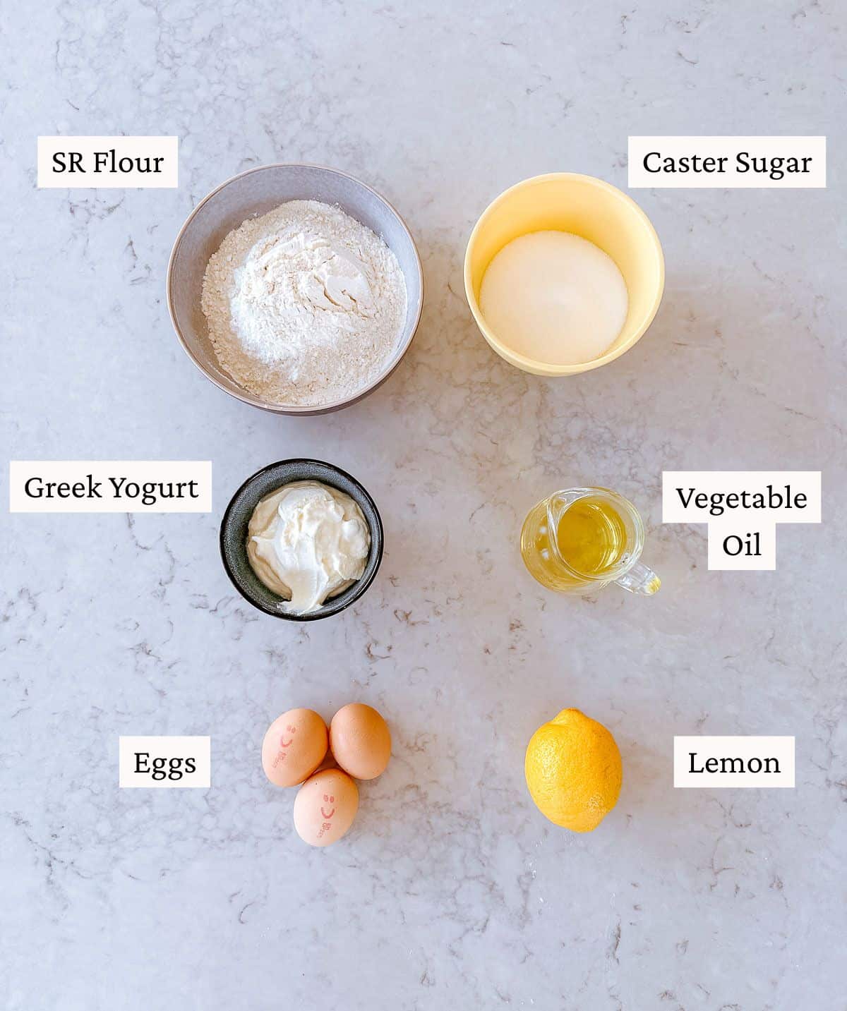 Six baking ingredients on a worktop: a bowl of self-raising flour, a bowl of caster sugar, a small bowl of Greek yoghurt, a small jug of vegetable oil, three eggs, and a whole lemon, each labelled.