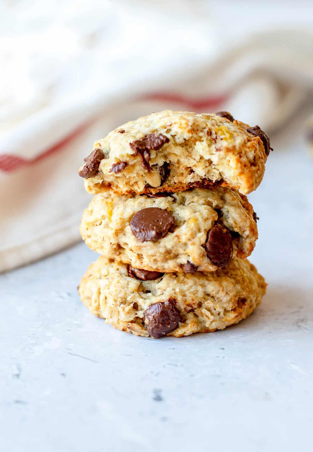 Three chocolate chip biscuits stacked on a light surface, with the top biscuit showing a bite taken out. A white cloth with a red stripe is blurred in the background.