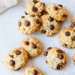Nine chocolate chip biscuits are scattered on a white surface, featuring golden brown edges and visible chocolate chips. A white cloth is partially visible in the upper left corner.