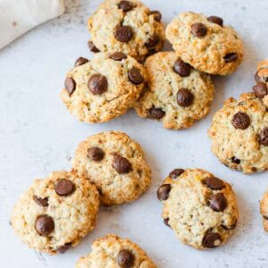 Nine chocolate chip biscuits are scattered on a white surface, featuring golden brown edges and visible chocolate chips. A white cloth is partially visible in the upper left corner.