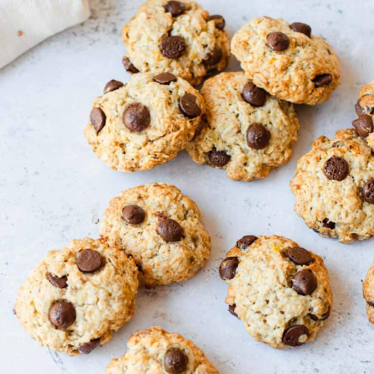 Nine chocolate chip biscuits are scattered on a white surface, featuring golden brown edges and visible chocolate chips. A white cloth is partially visible in the upper left corner.