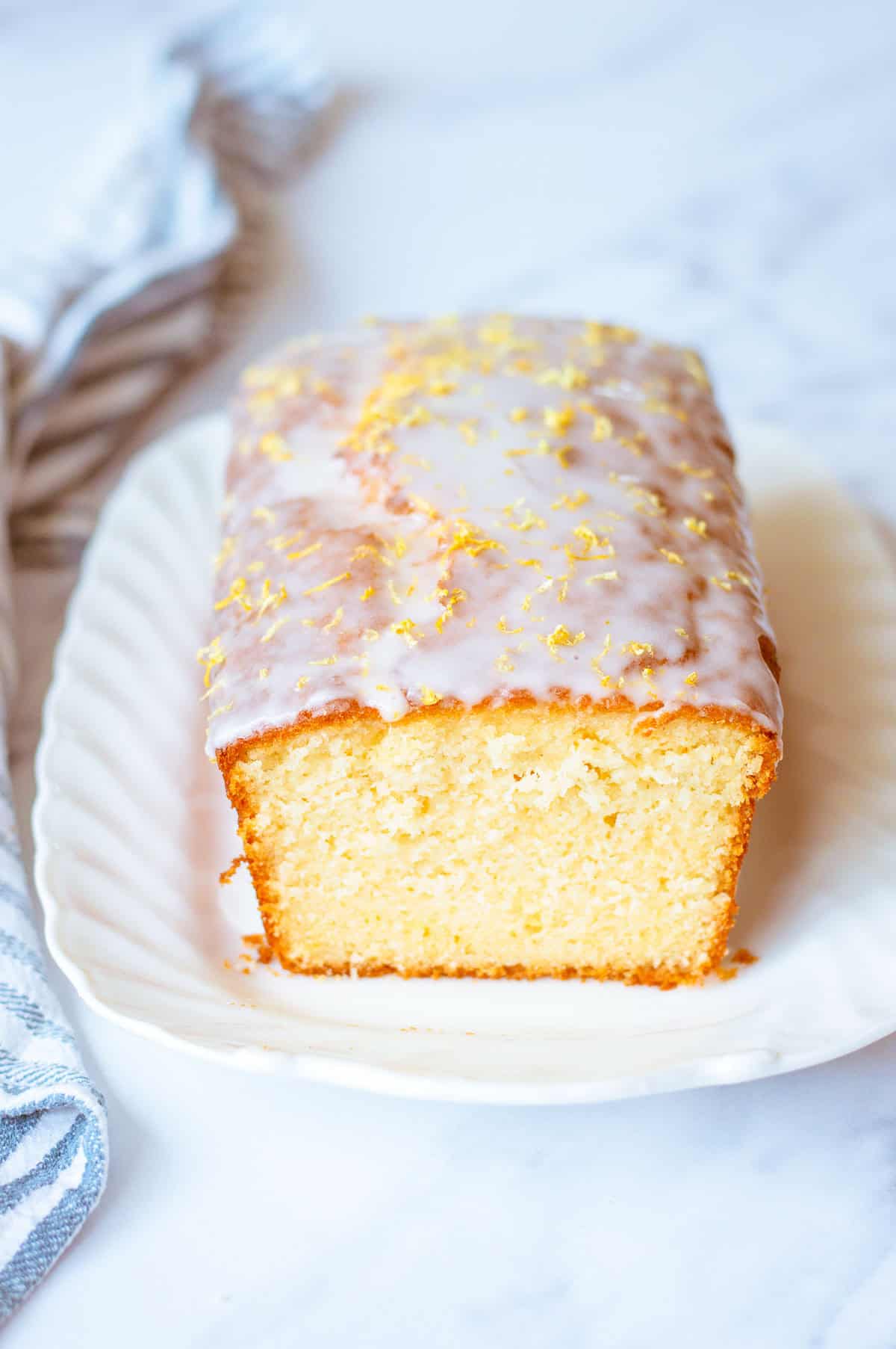 A loaf of glazed lemon cake sits on a white plate. The cake is topped with icing and lemon zest, and one end has been sliced to reveal the soft, moist interior.