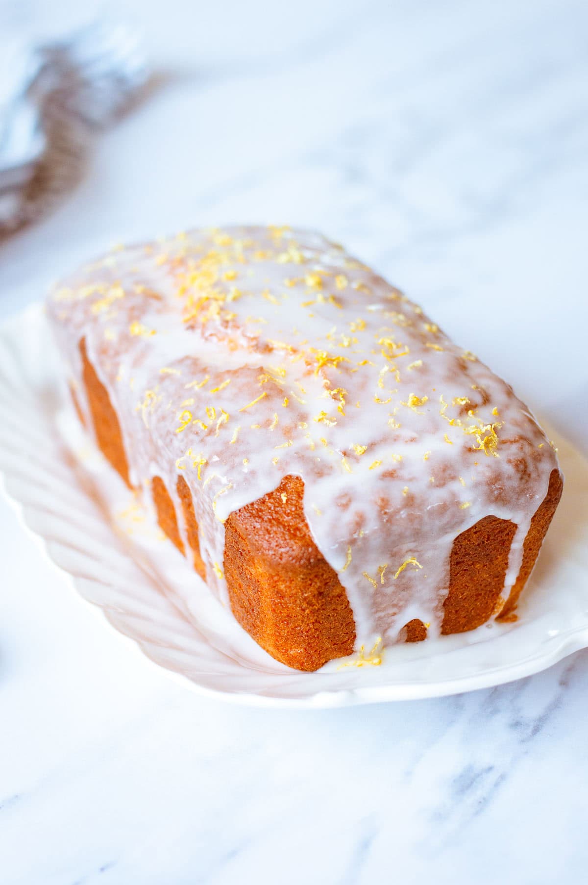 A loaf of lemon cake on a white plate, topped with white icing and sprinkled with lemon zest, set on a marble surface.