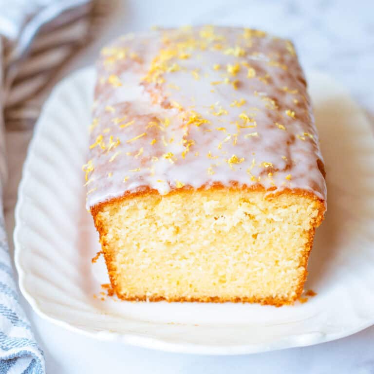 A loaf of glazed lemon cake sits on a white plate. The cake is topped with icing and lemon zest, and one end has been sliced to reveal the soft, moist interior.