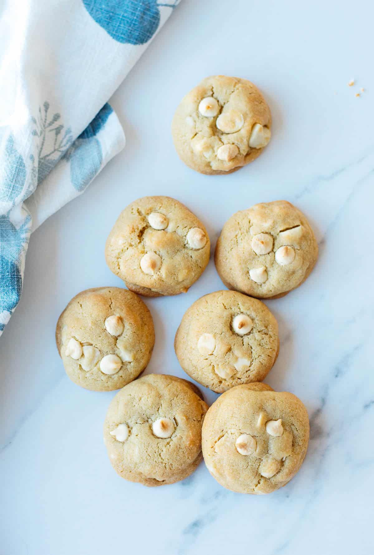 Seven golden-brown biscuits with white chocolate chips sit on a white marble surface next to a blue and white cloth.