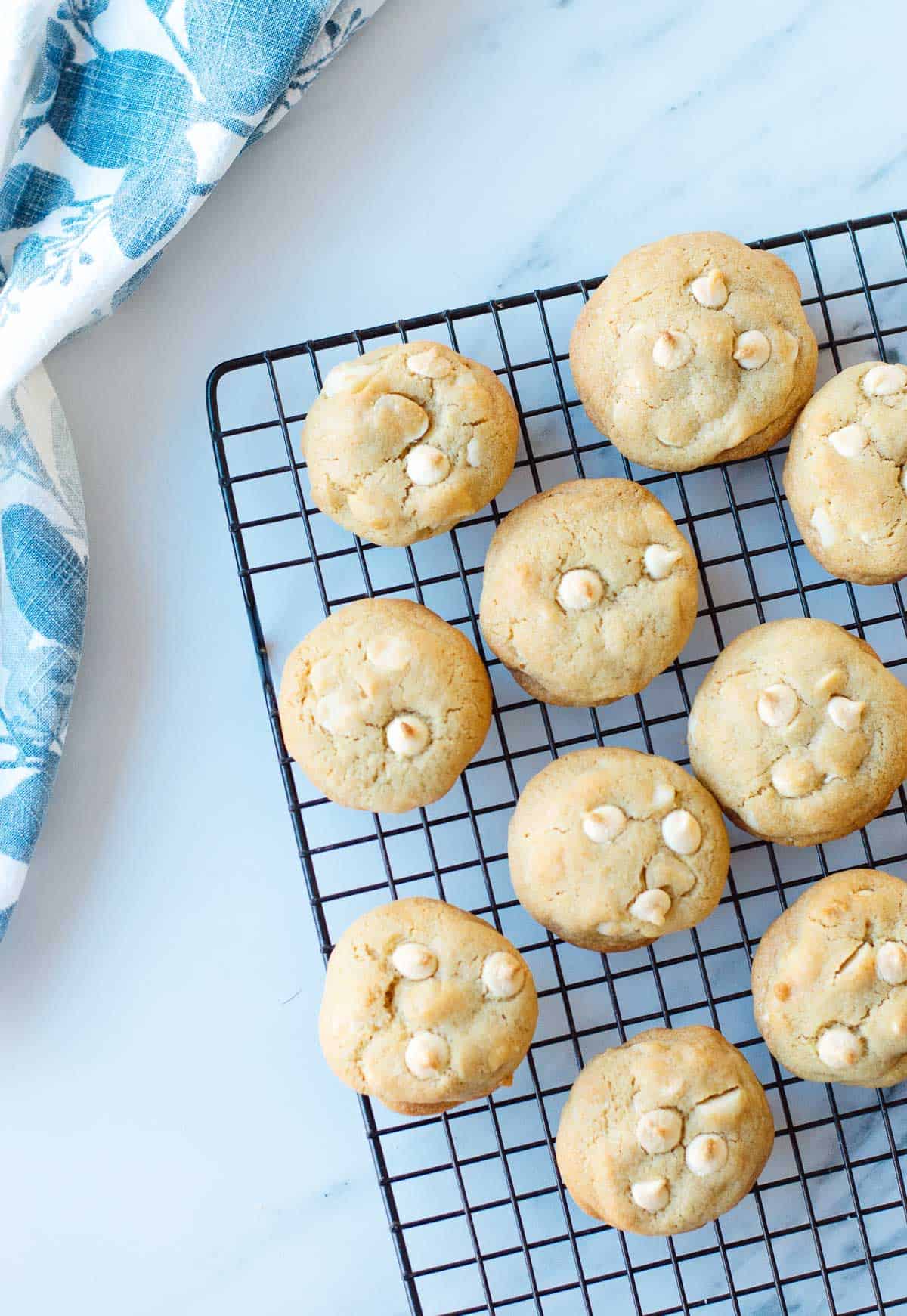 White chocolate chip biscuits cool on a black wire rack atop a white marble surface, with part of a blue and white cloth visible in the corner.