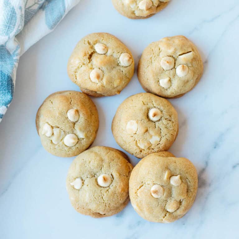 Six golden-brown biscuits with white chocolate chips sit on a white marble surface next to a blue and white cloth.