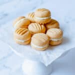 A stack of round, sandwich-style biscuits with cream filling, arranged on white baking parchment atop a white cake stand against a light background.